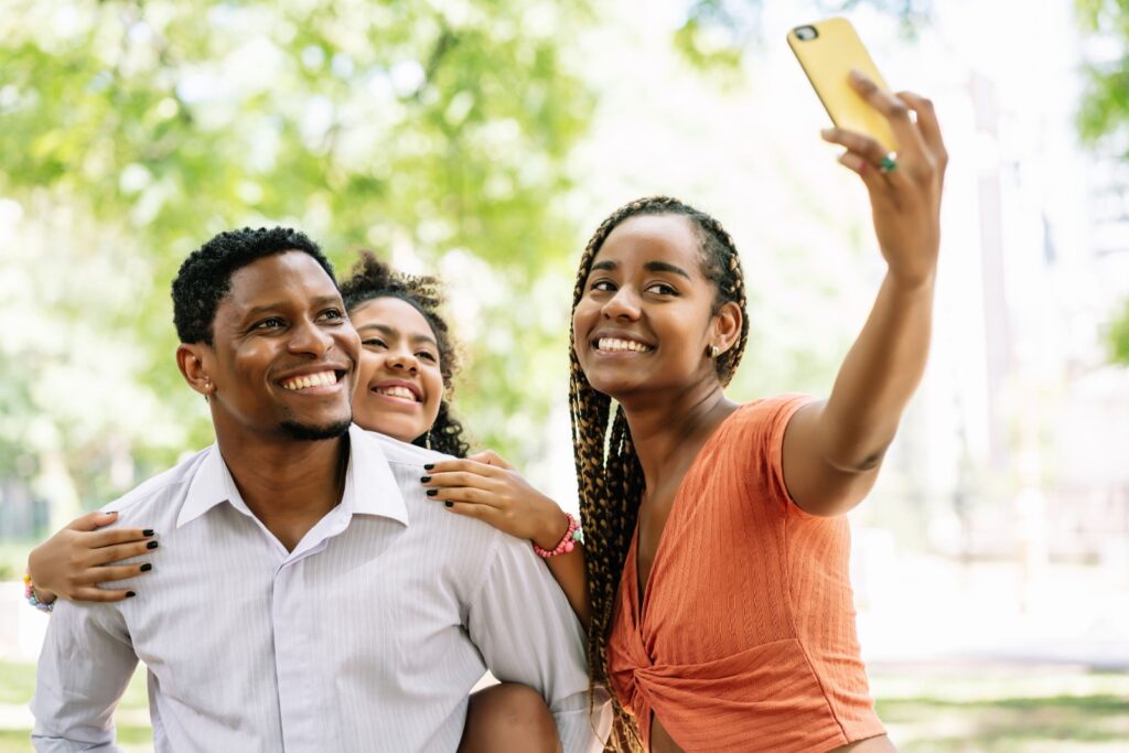 Três jovens negros sorrindo enquanto tiram uma selfie em um parque arborizado, com um homem ao centro e duas mulheres ao redor.