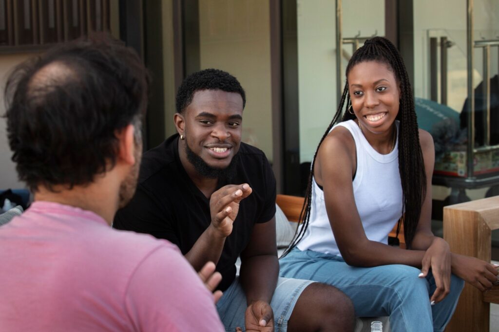 Dois jovens negros conversando e sorrindo com um amigo em um espaço ao ar livre, criando um ambiente acolhedor e amigável.